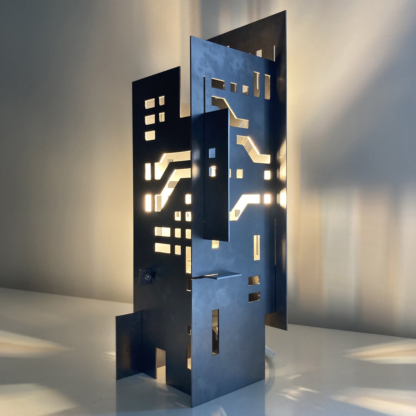 Modern steel cityscape table lamp with layered architectural panels and geometric cutouts casting warm ambient light and shadows.
