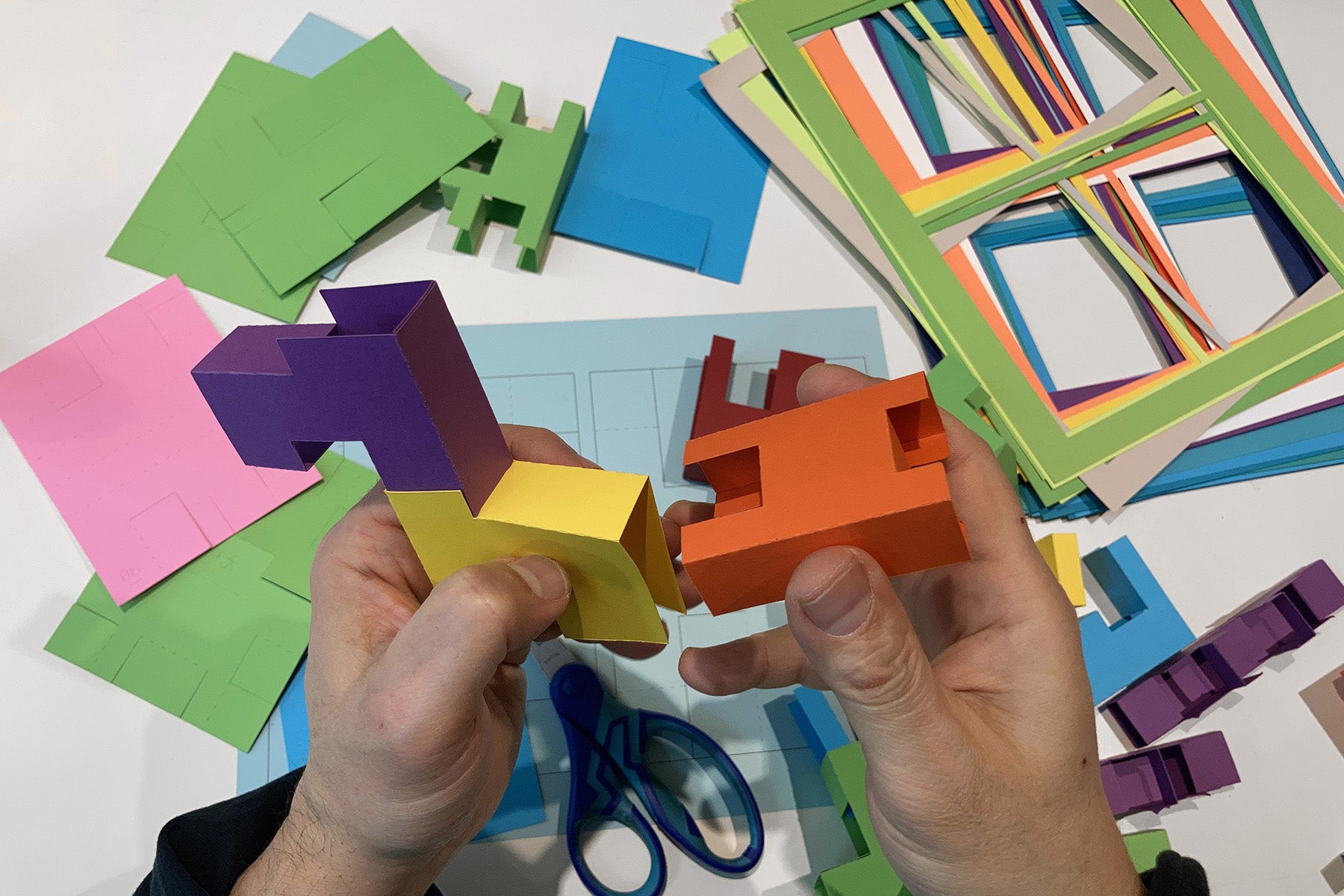 Colorful modular paper shapes and scissors on a white surface with hands assembling them to build an animal