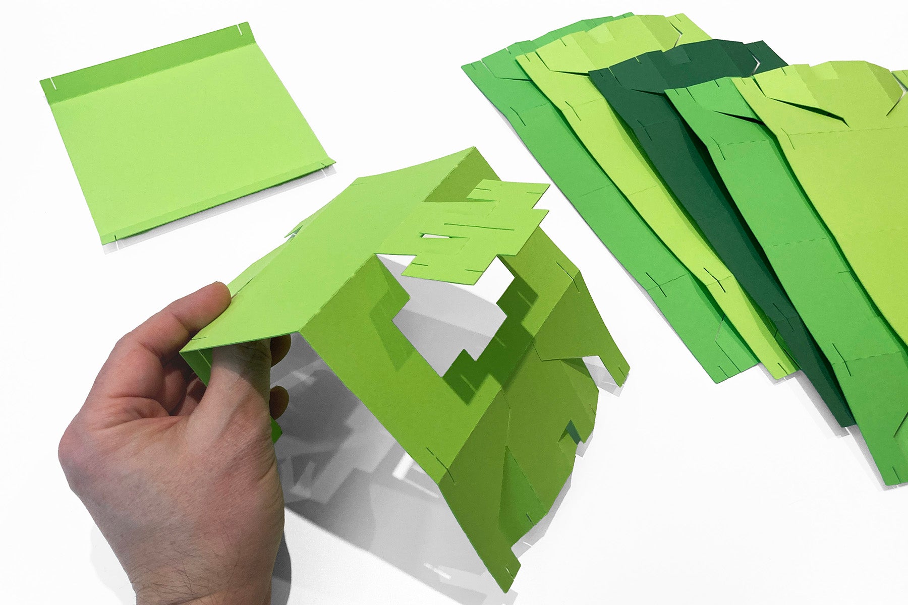 Hand assembling a green paper craft with additional green paper sheets on a white background