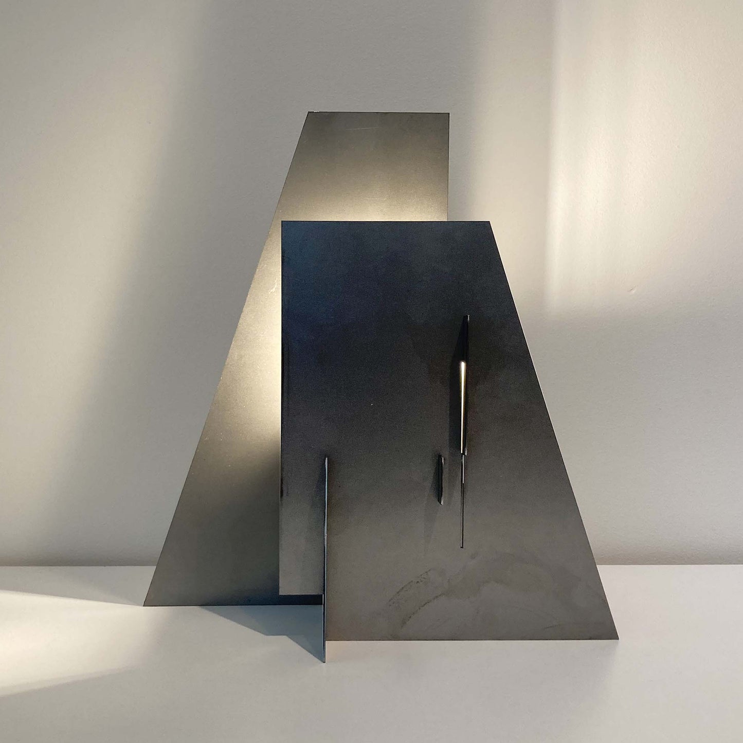Illuminated modern metal sculptural lamp casting a shadow on a plain wall.