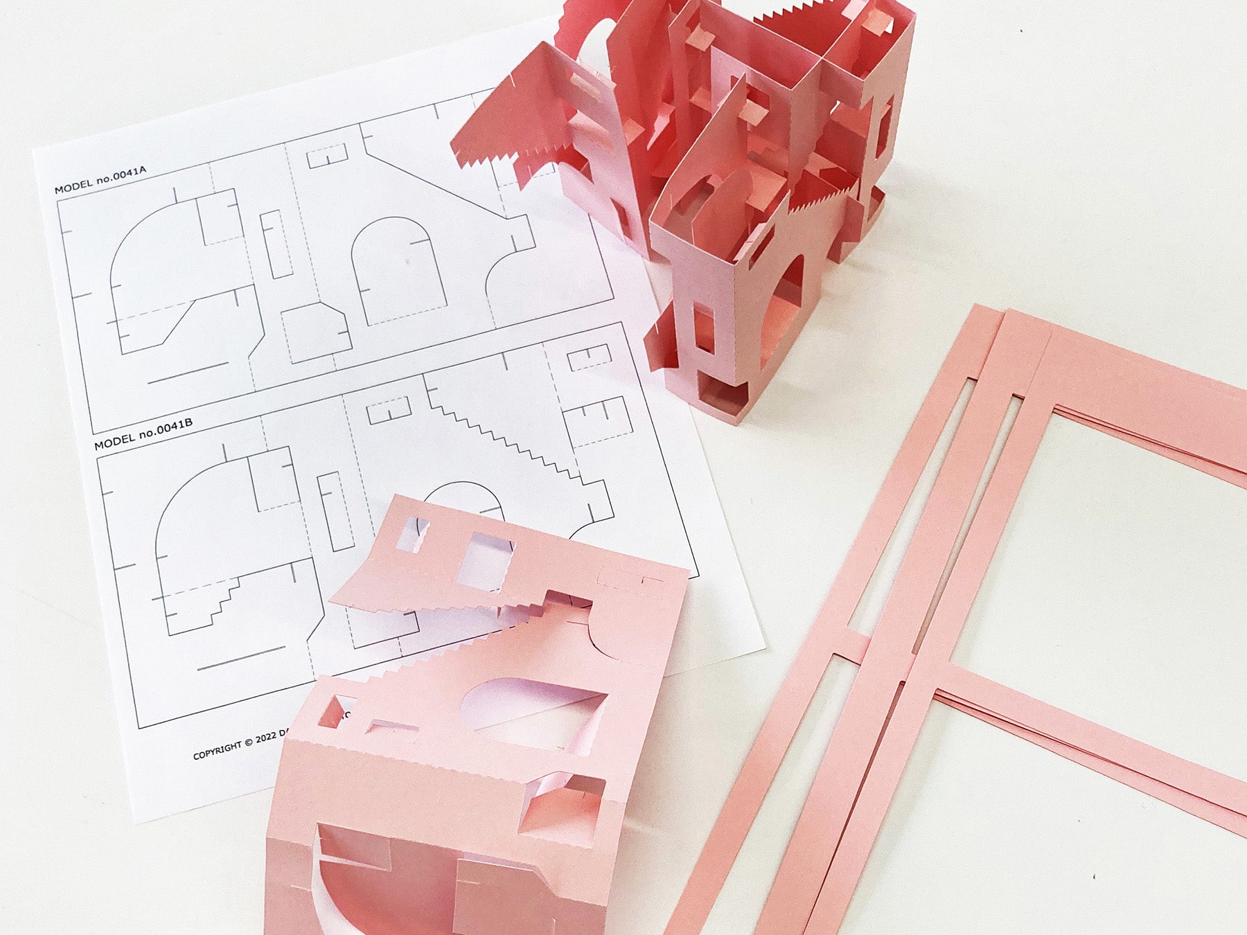 Pink architectural paper model kit with building pieces and a design sheet on a white background